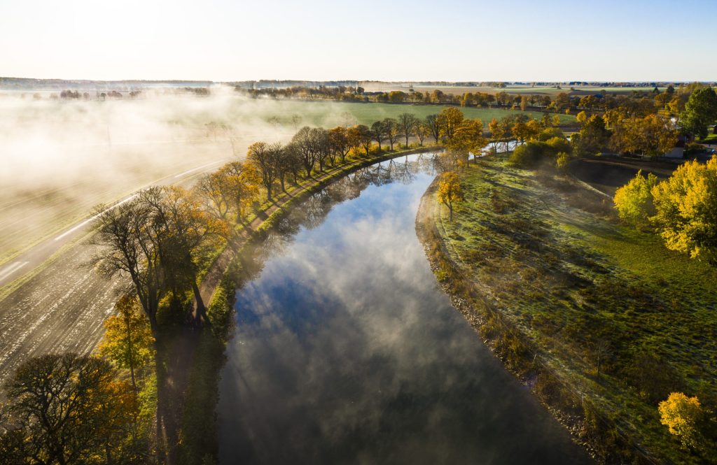 Flygbild över Göta kanal en dimmig morgon med höstfärgade träd längs kanalbanken och landskapet som breder ut sig i bakgrunden.